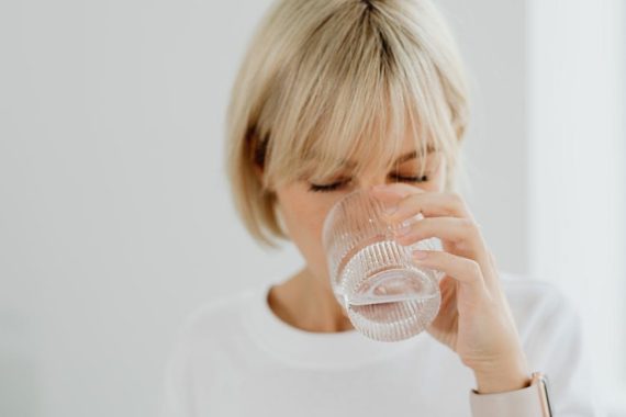 Blonde woman drinking water from glass indoors, showcasing healthy lifestyle and hydration.